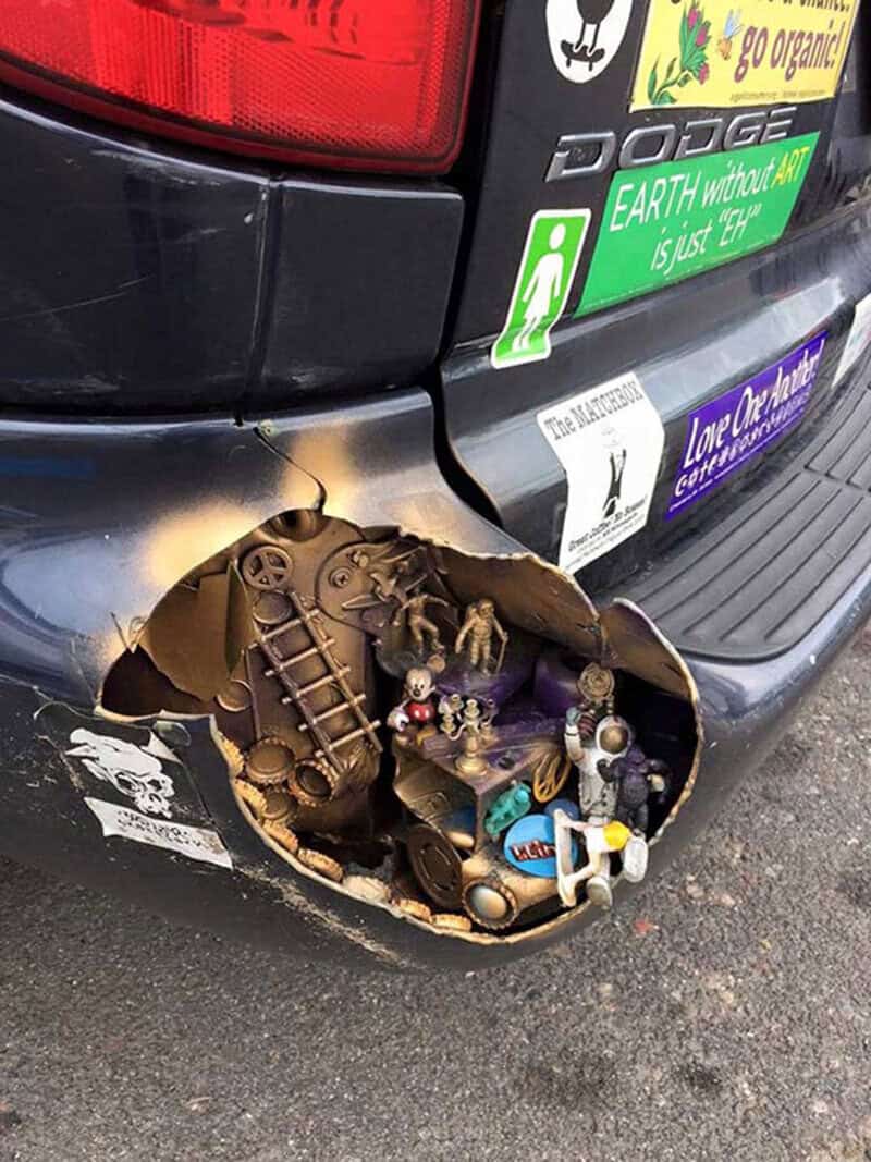 Broken car bumper with a miniature scene inside the hole, including small figurines and toys.
