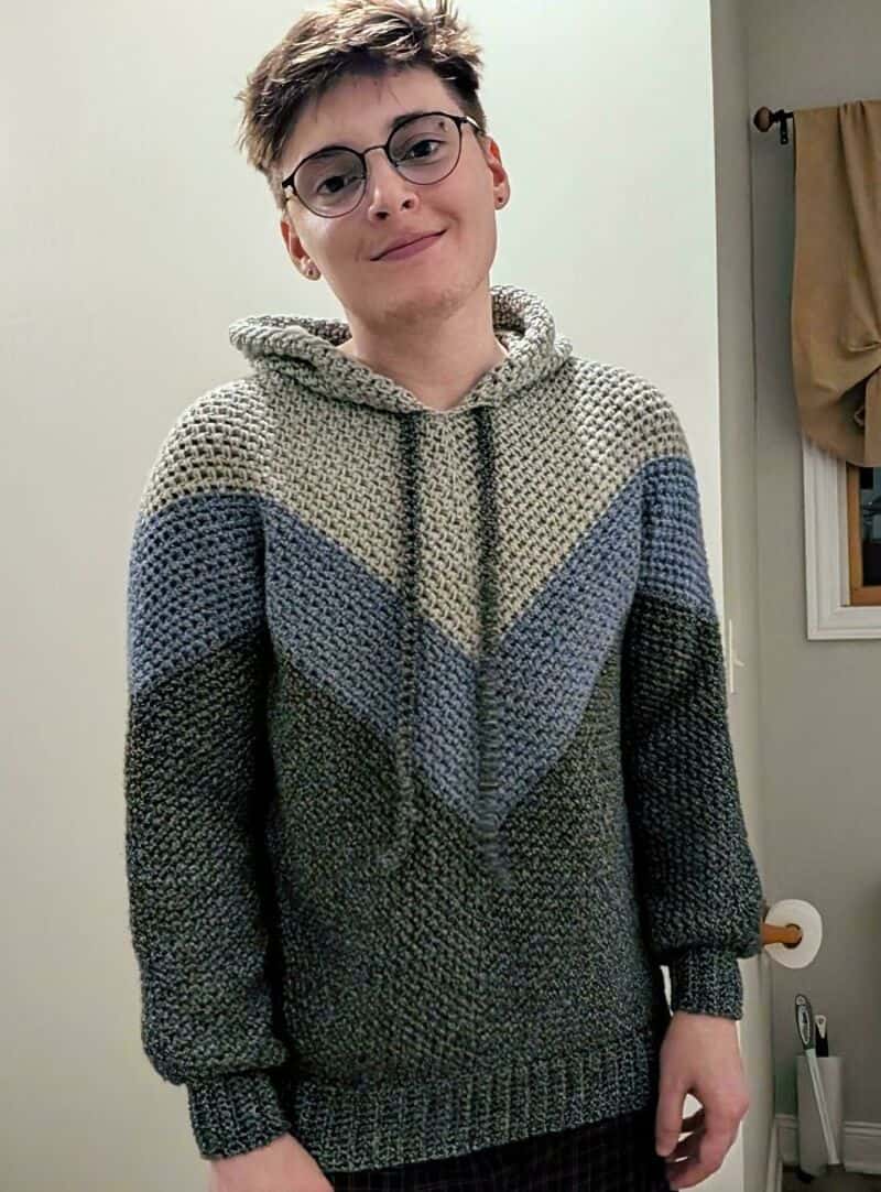Young person with short hair and glasses wearing a multicolored crochet hoodie.