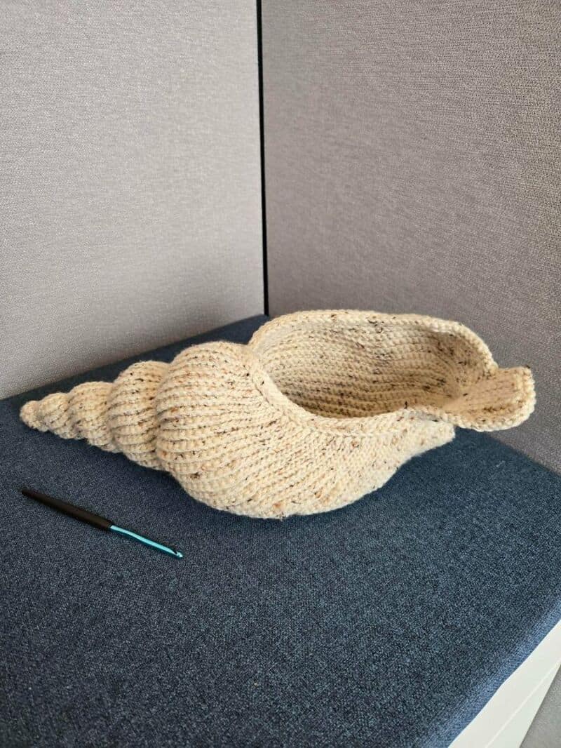 Cream-colored crochet seashell hat on a blue surface with a crochet hook nearby.