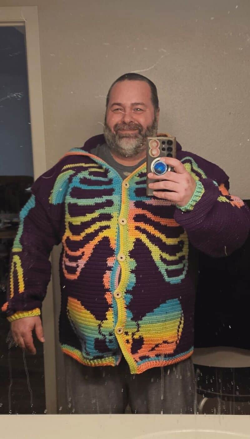 Man taking mirror selfie wearing a colorful, rainbow crochet cardigan.
