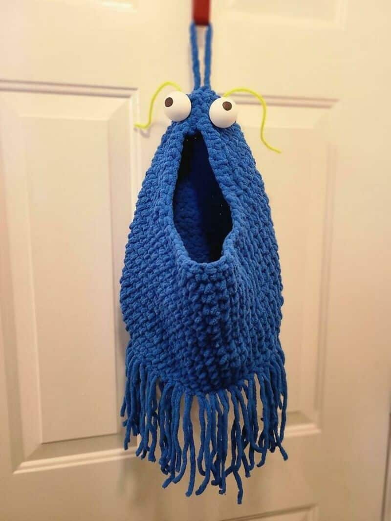 Blue crochet bag with googly eyes and yellow antennae hanging on door.