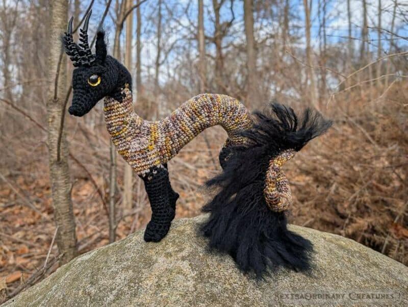 Handmade crochet dragon with black horns, tail, and limbs, on a rock in a forest.