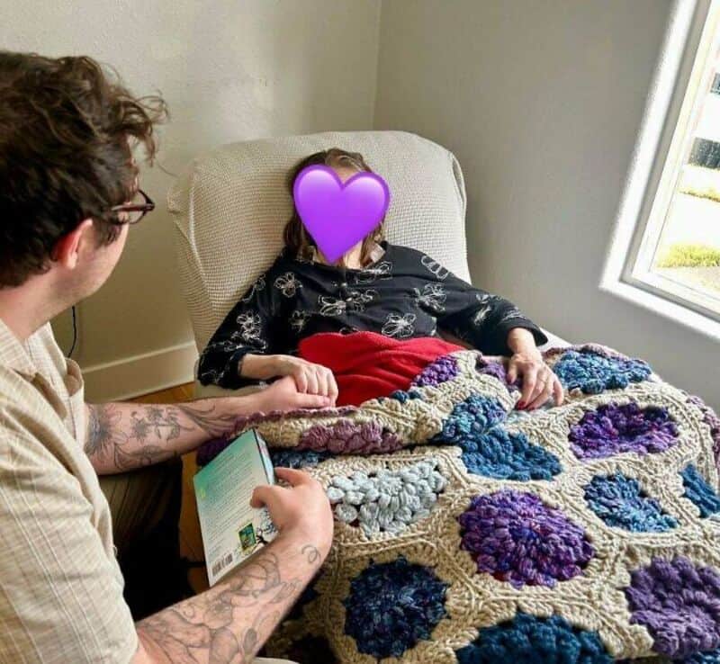 Person with tattoos reading to a woman covered with a colorful crochet blanket.
