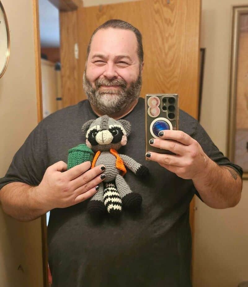 Man taking a selfie with a crocheted raccoon doll in a mirror.