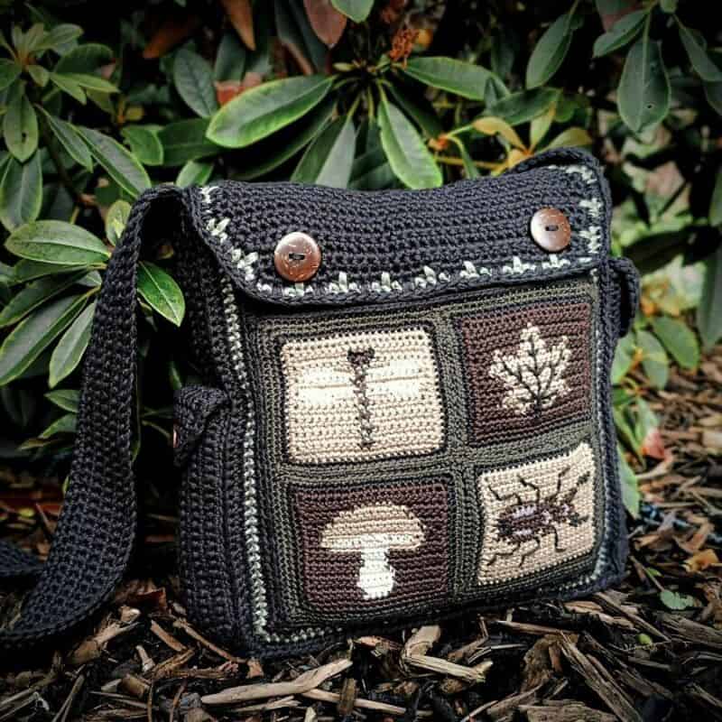 Crochet bag with nature-themed patches, black strap, and button details, outdoors among leaves.