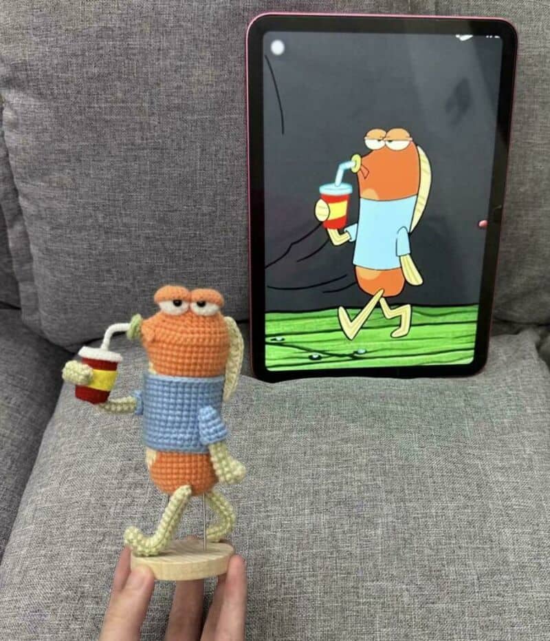 Crochet character holding a drink with a cartoon image on a tablet behind it.