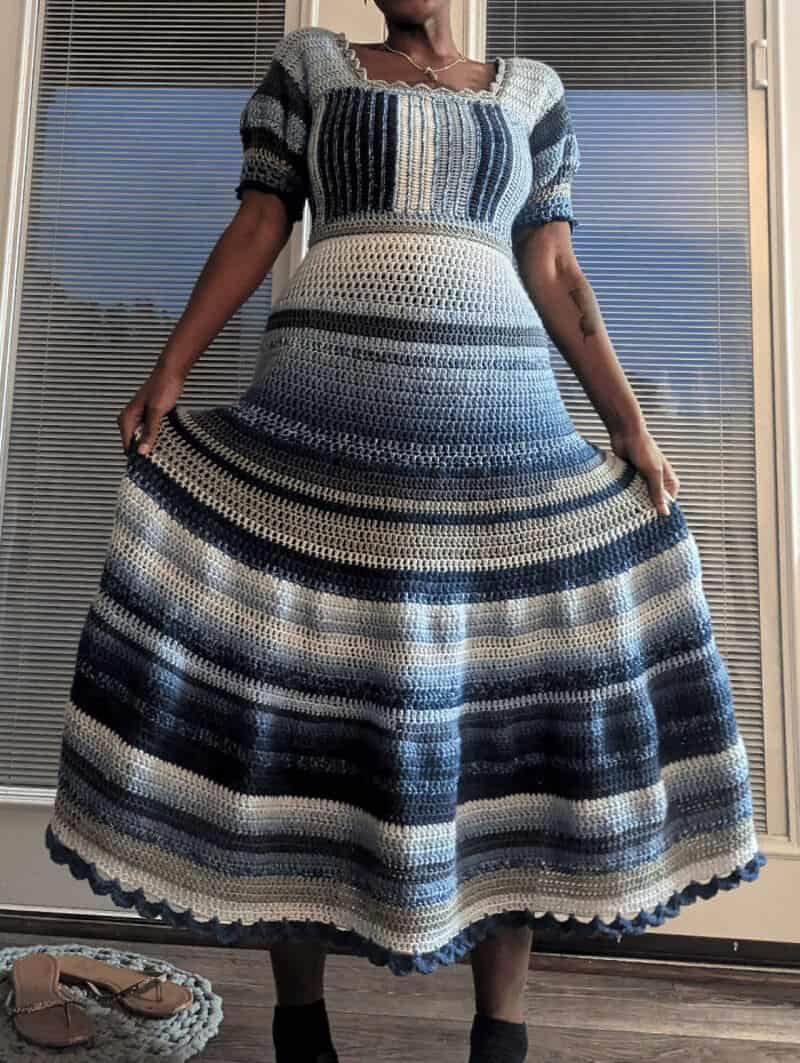 Woman in a striped crochet dress holding the sides, standing in front of blinds.