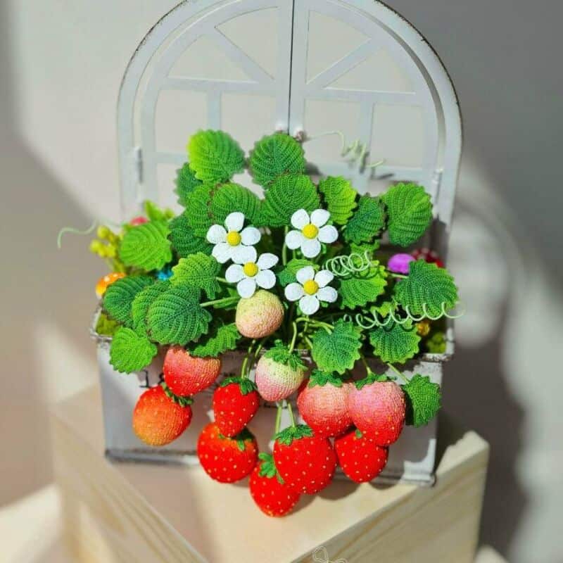 Colorful crochet strawberry and flower arrangement in a white basket.