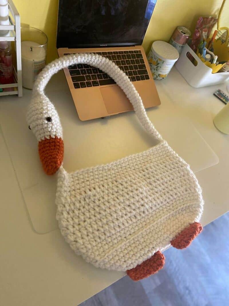 Crochet swan bag with orange beak and feet on a desk with laptop and stationery.
