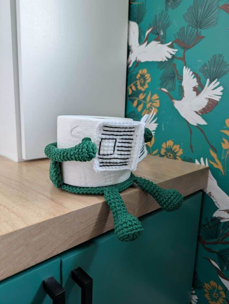 Crochet robot with white body, green arms and legs, on a wooden surface.