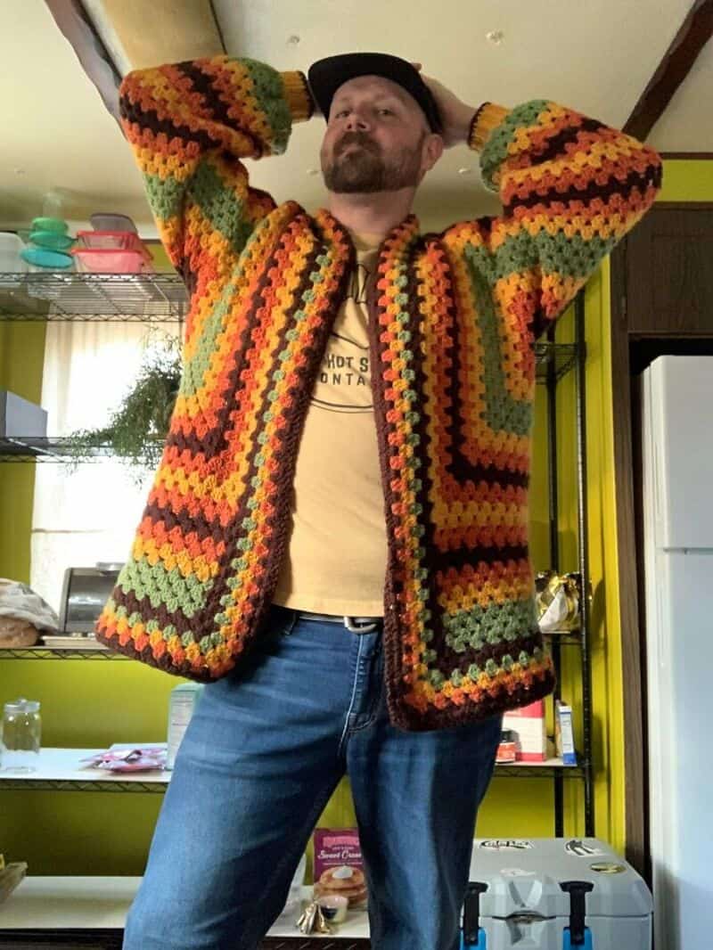 Man wearing a colorful crochet jacket with arms raised in a kitchen.