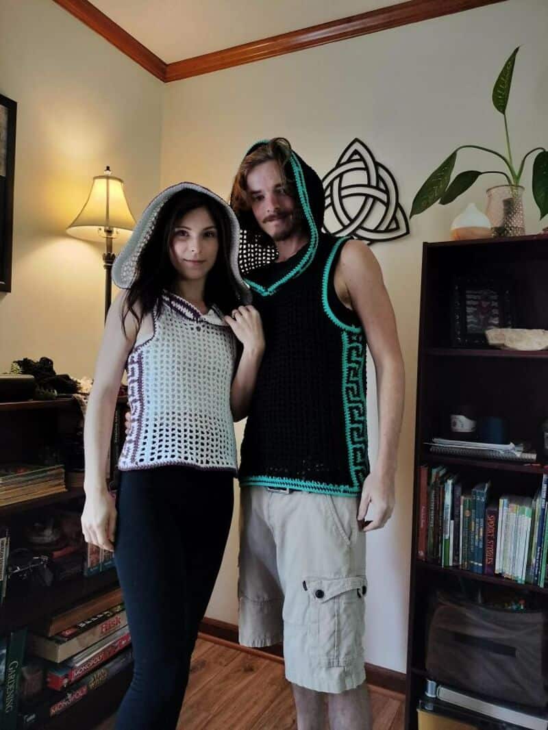Two people in crochet outfits stand in a room with a bookshelf and lamp.