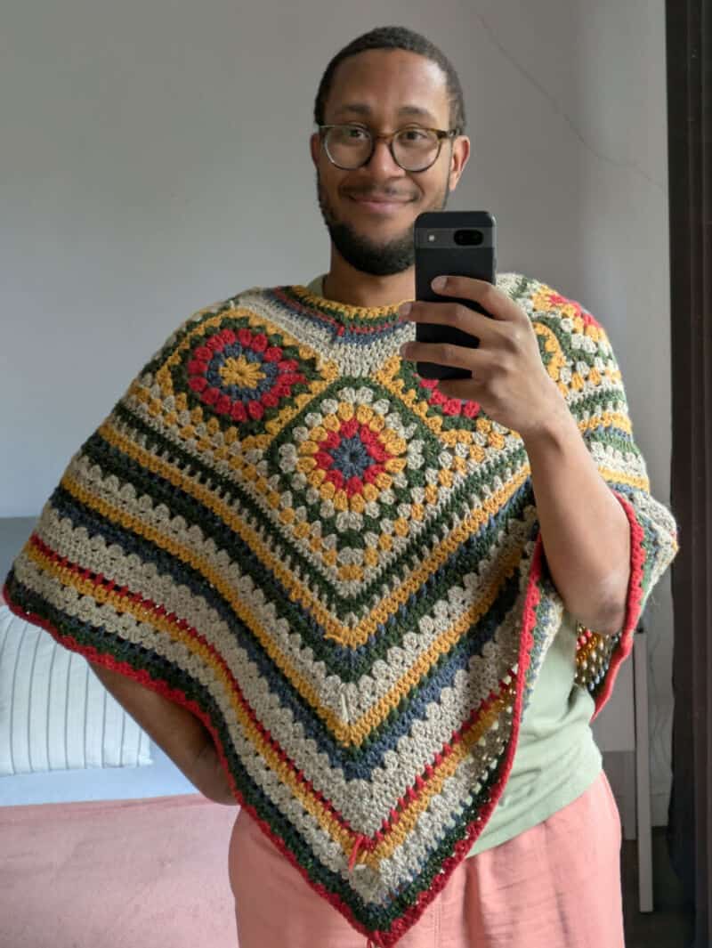 Smiling man taking a selfie in a crochet poncho with colorful granny squares.