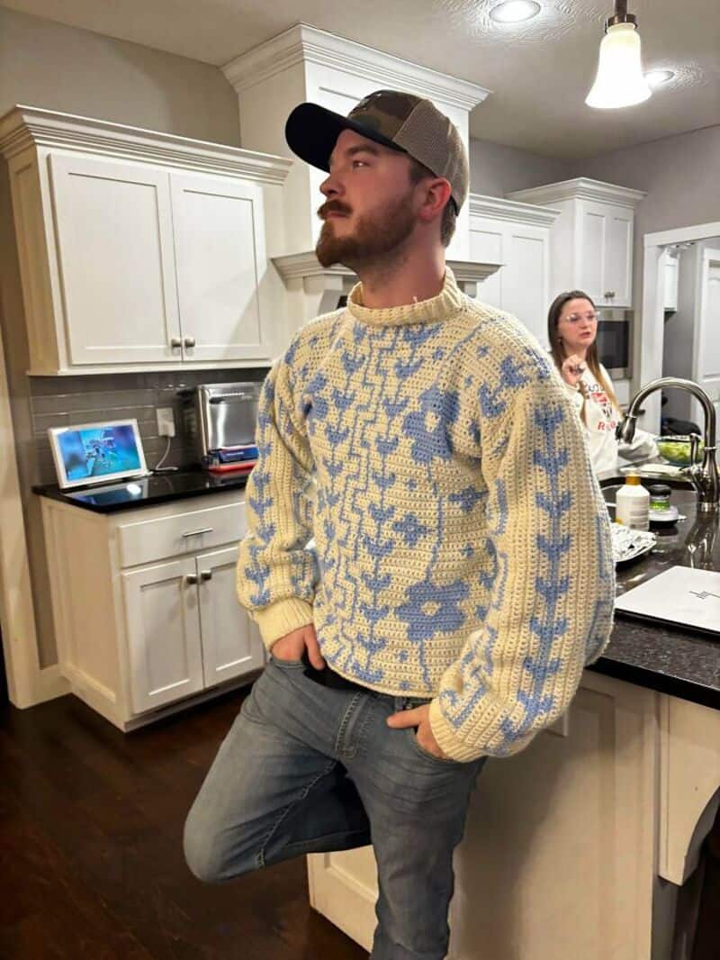 Man in a crochet sweater in a kitchen with a woman in the background.