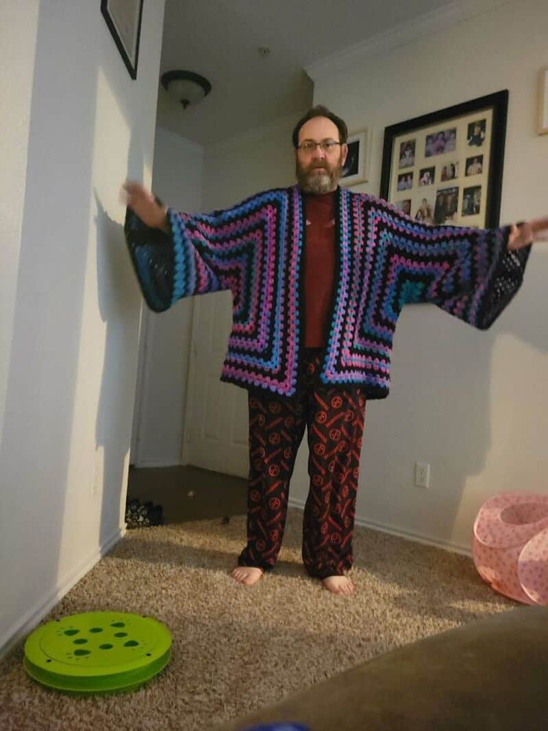 Person in colorful crochet shawl with arms outstretched in a room.