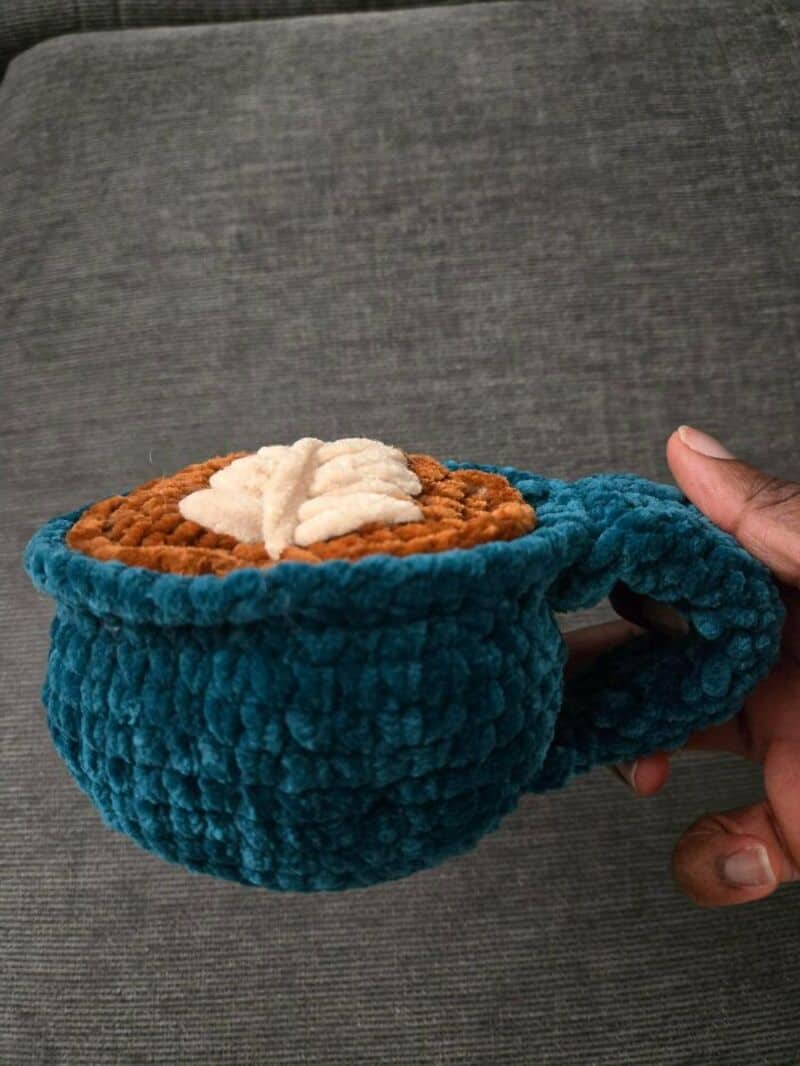 Hand holding a blue crochet mug with a brown and white crochet design inside.