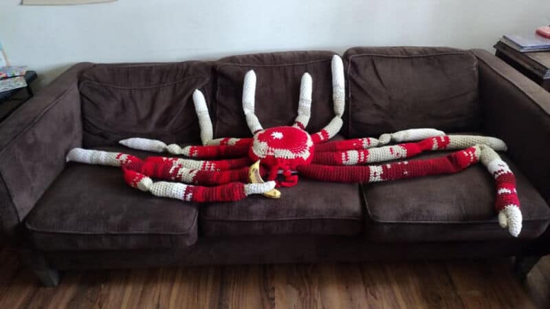 Large crocheted spider with red and white body and long legs on a brown sofa.