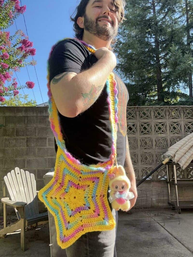Man with a crochet bag and plush toy in backyard with trees and blue sky.