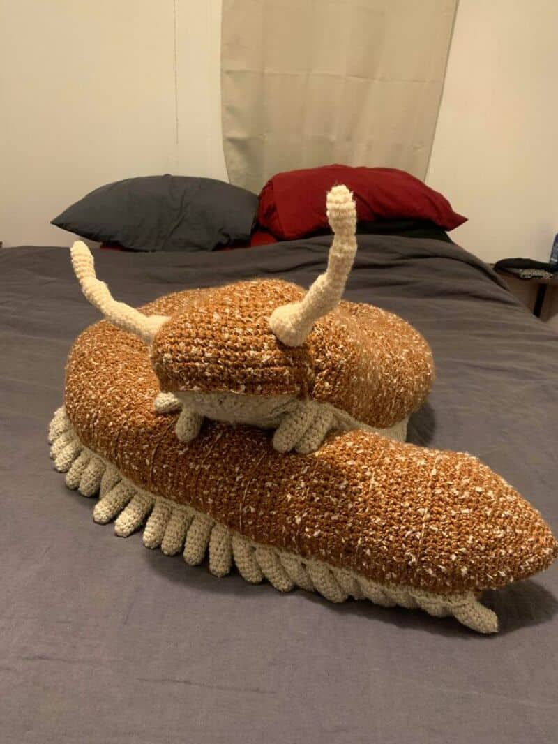 Crocheted crab with antennae and claws on a bed with pillows.