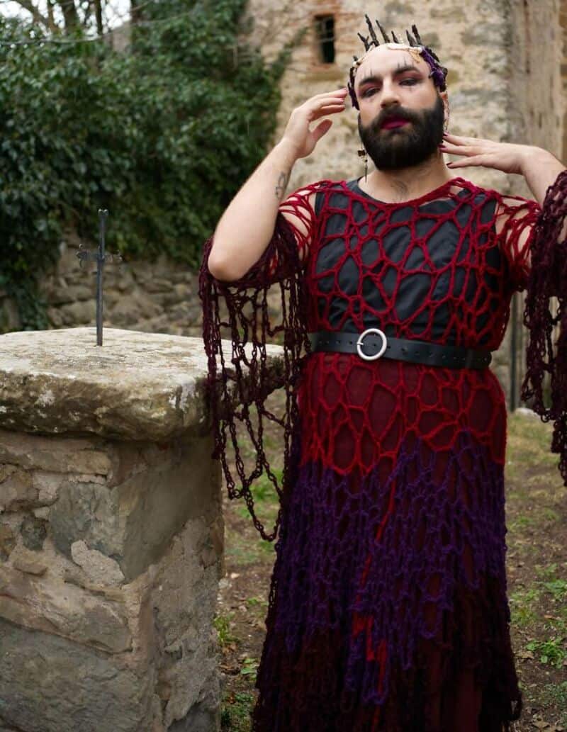 Person with makeup and a crochet dress outdoors near a stone wall.