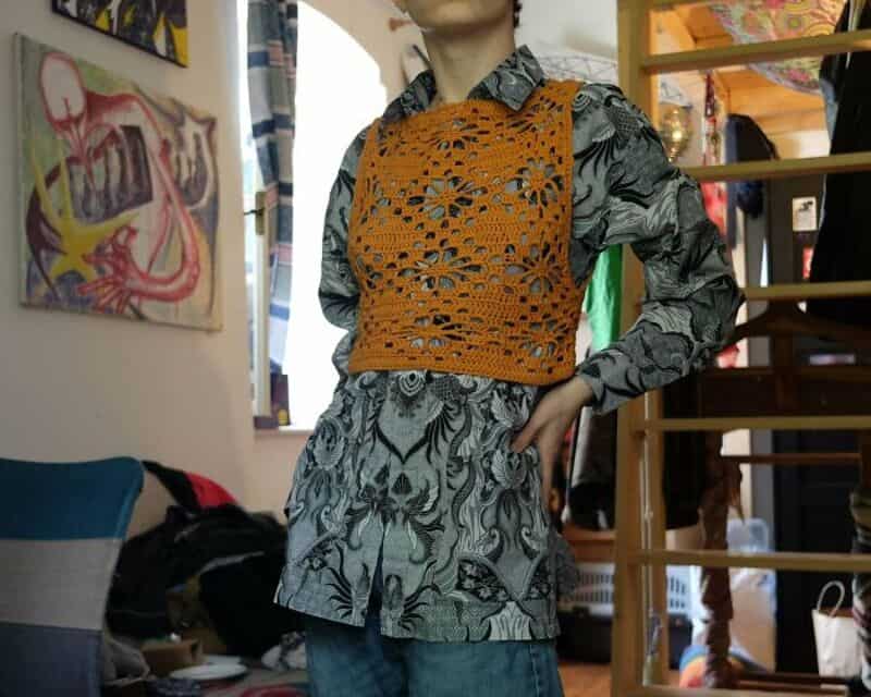 Person wearing a patterned shirt with an orange crochet vest in a colorful room.