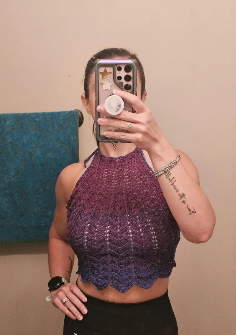 Person taking a mirror selfie wearing a purple crochet top, holding phone with star stickers.