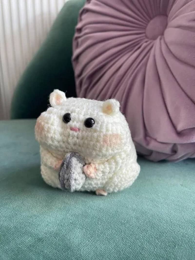 Cute crocheted hamster holding a gray object, with a purple pillow and green cushion in background.