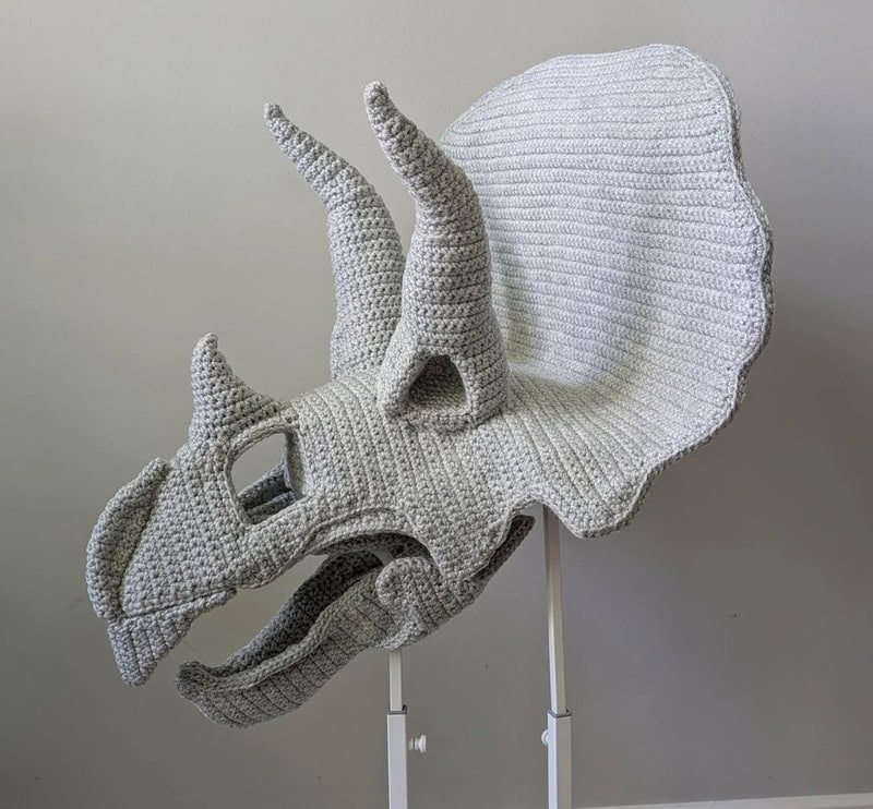 Crochet triceratops sculpture with a large frill and horns on a stand.