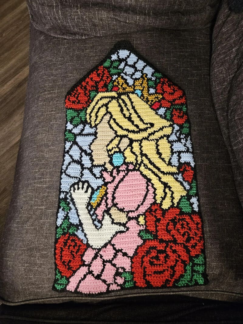 Colorful crochet art of a girl with roses and blonde hair on a gray couch.