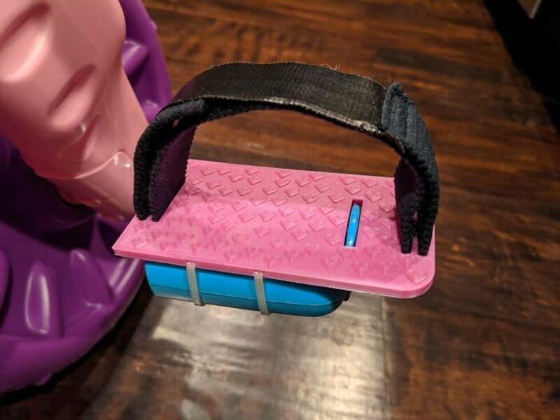 Pink skateboard with a black strap and blue wheels on a wooden floor.