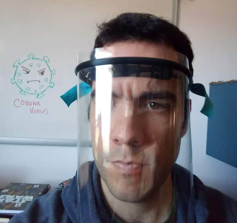 Man wearing face shield in front of whiteboard with coronavirus drawing and text.