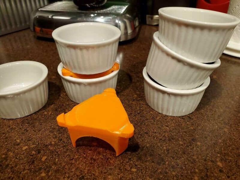 Stacked white ramekins and an orange 3D-printed object on a brown countertop.