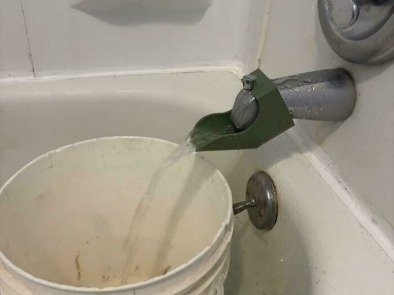 Faucet with green cover pouring water into a white bucket in a bathroom.