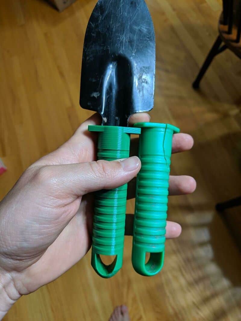 Hand holding a small gardening trowel and a hand rake with green handles.