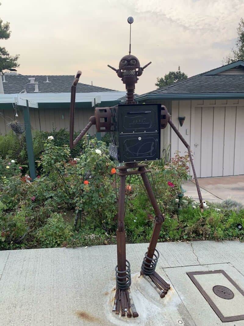 Rusty robot sculpture with antenna, arms, and legs in a garden.