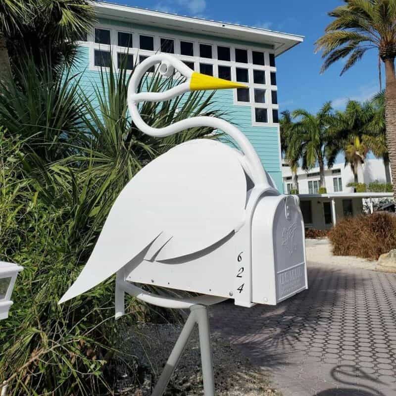 White mailbox shaped like a heron bird with a yellow beak, in front of a blue house.