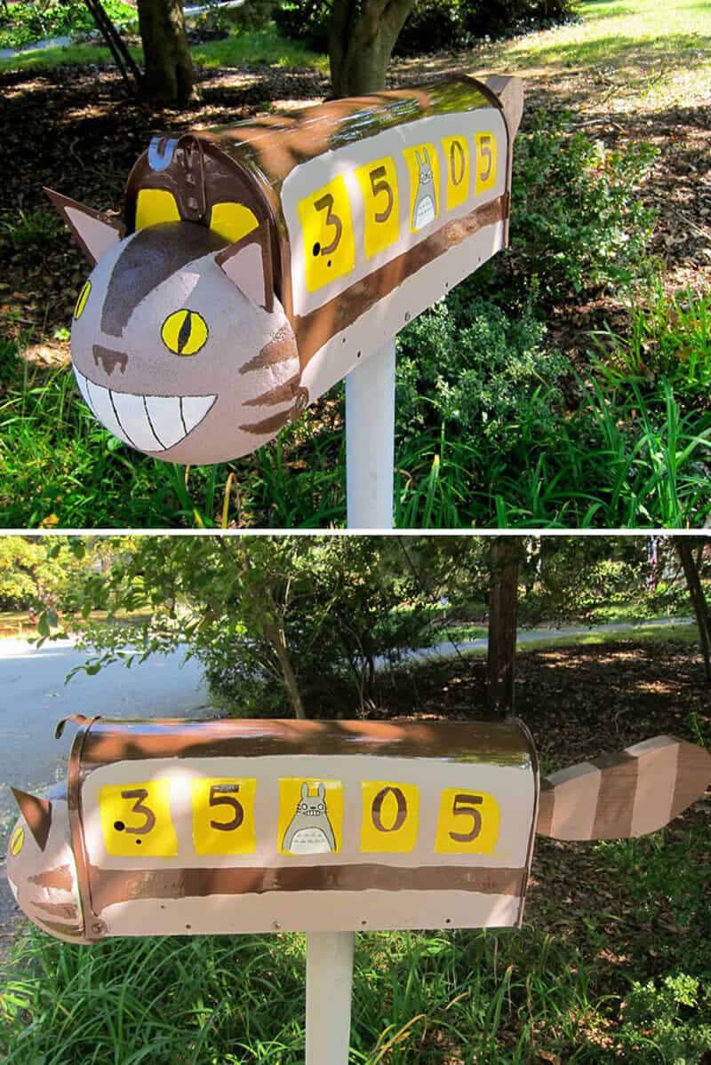Cat-shaped mailbox with yellow number blocks and a tail, in a park setting.