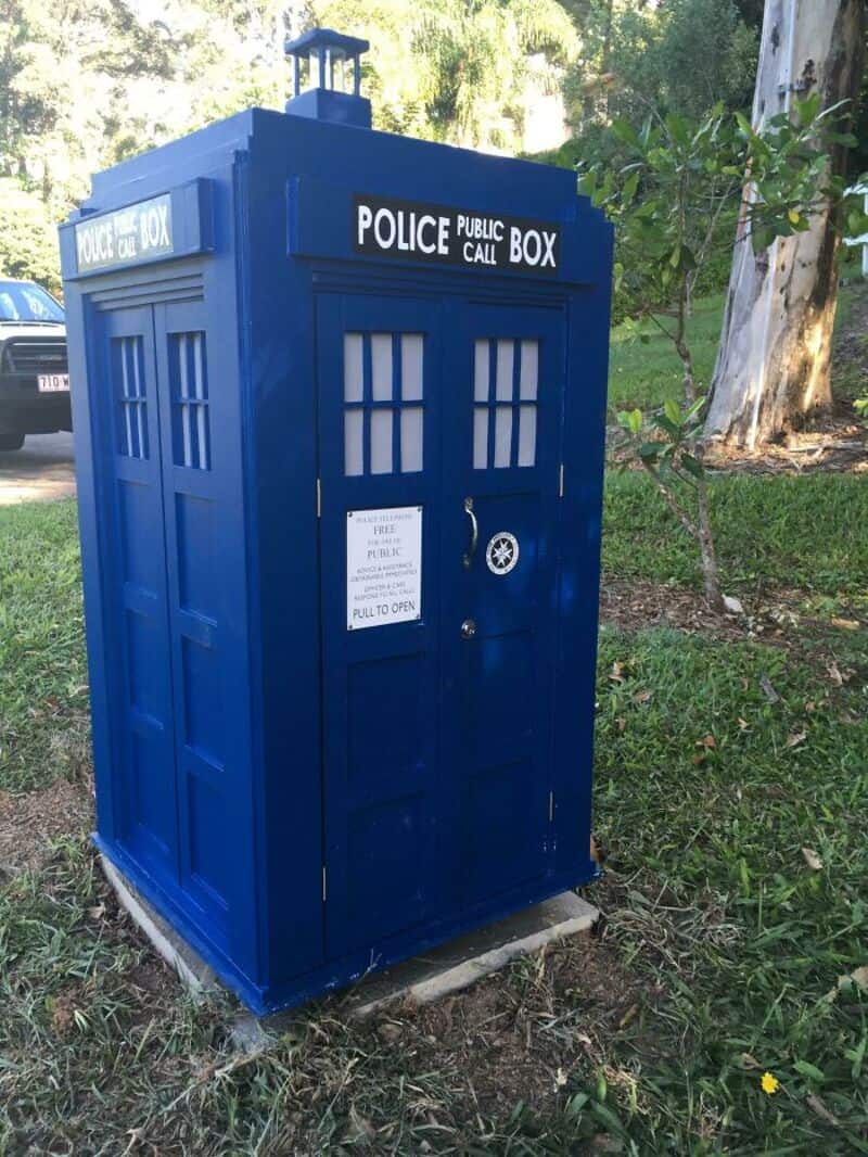 Blue police public call box with windows and sign, outdoors near trees.