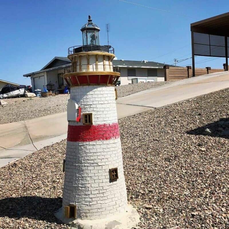 Miniature lighthouse with mailbox and small windows, on a gravel surface.