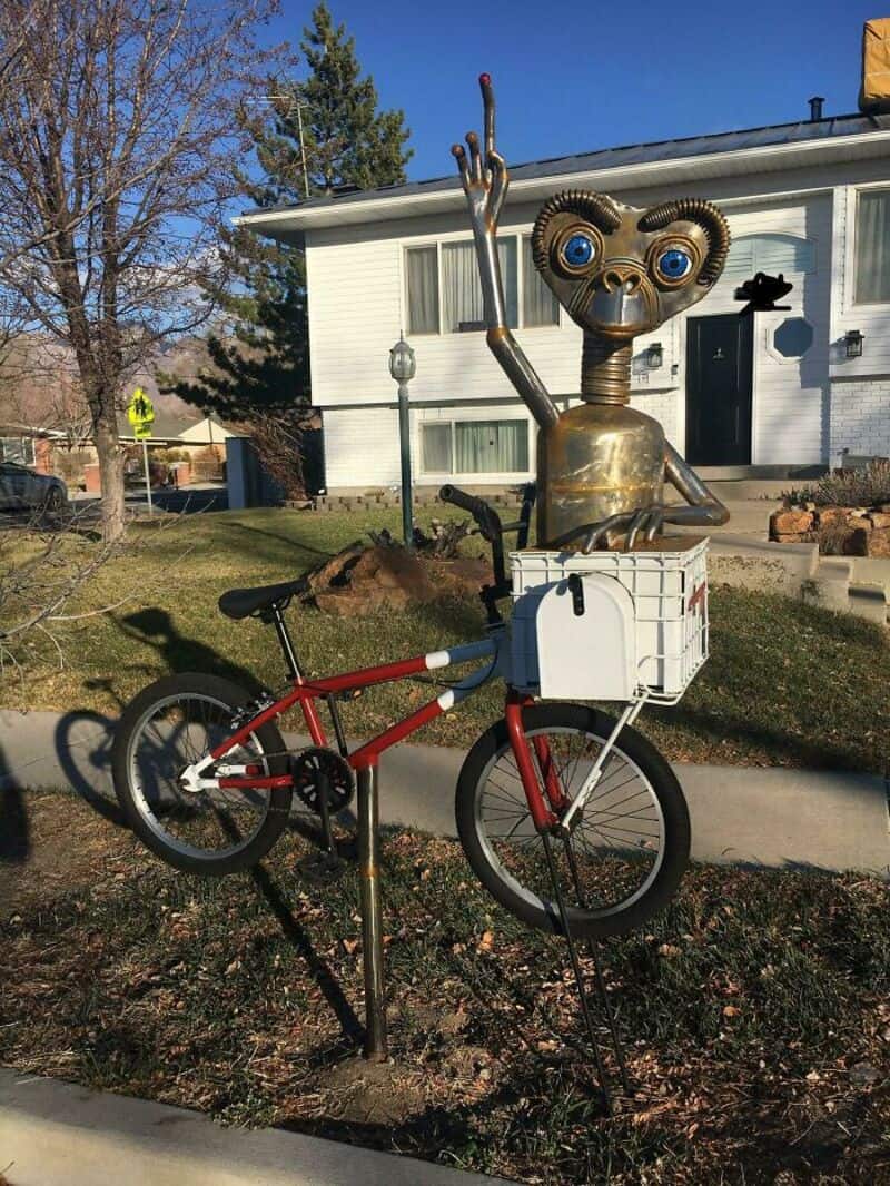 Sculpture of a robot with a monkey face on a bicycle in front of a house.
