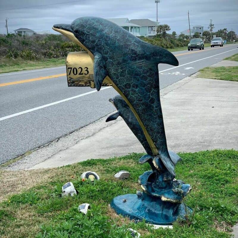 Dolphin-shaped mailbox with the number 7023 on it, on a grassy area near the road.