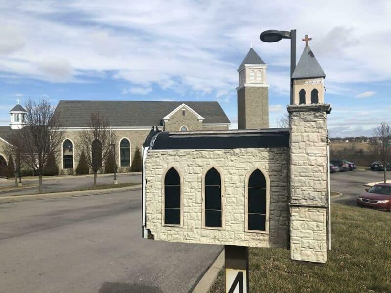 A church-shaped mailbox with a cross on top, set in a parking lot.