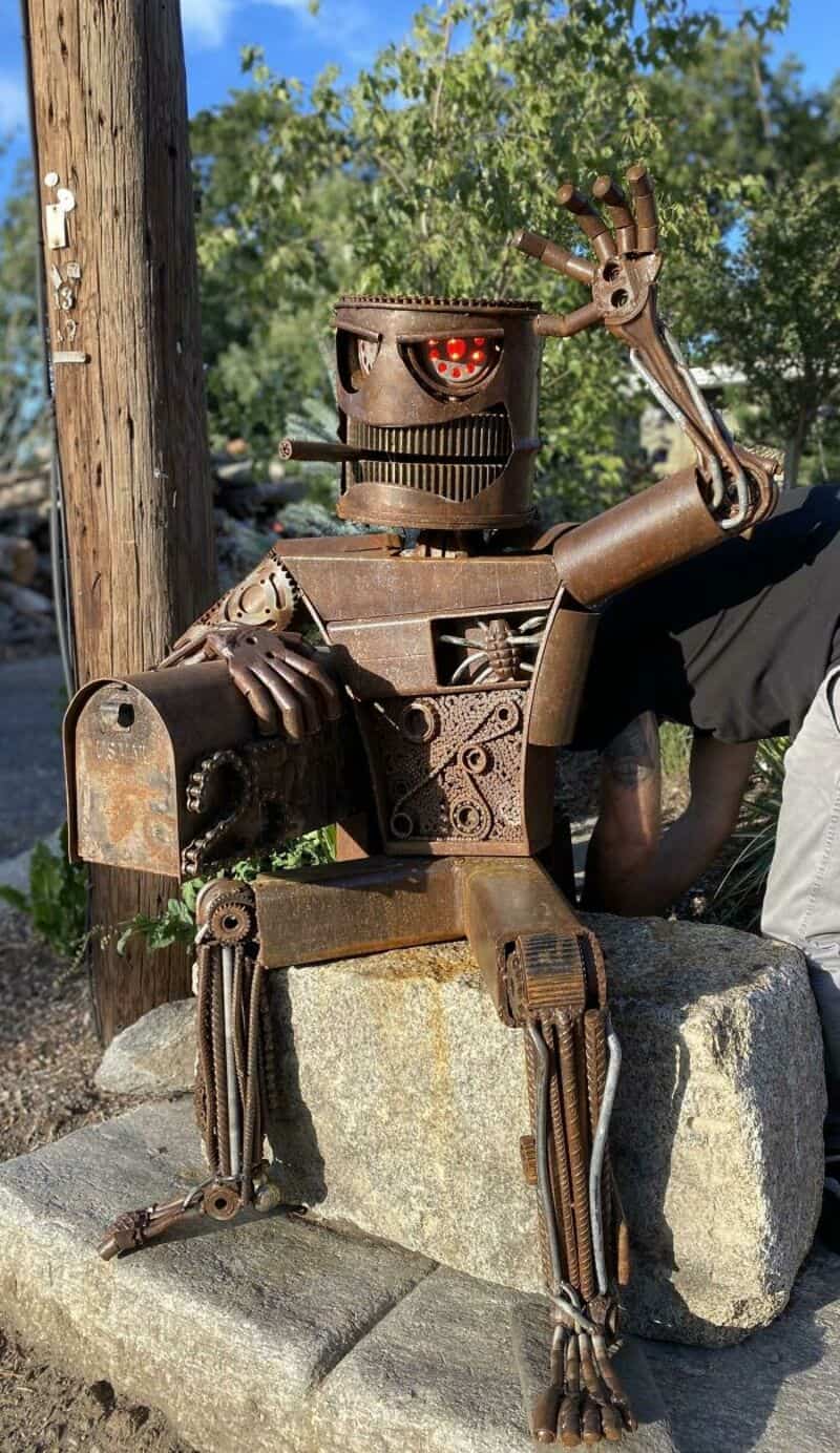 Rusty robot sculpture sitting on a stone with a hand raised in a wave.