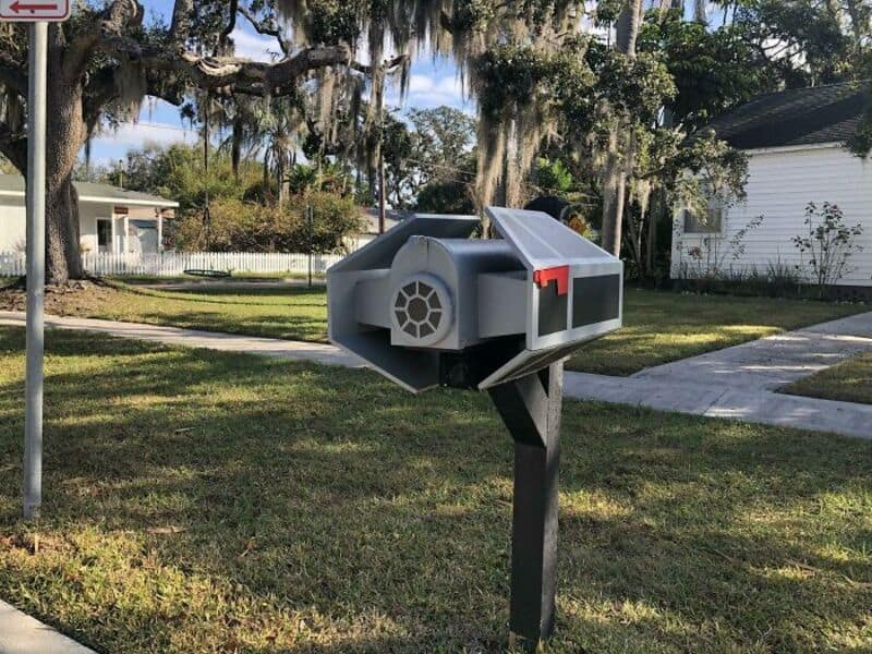 A gray mailbox with a red flag on a black post in a grassy yard.