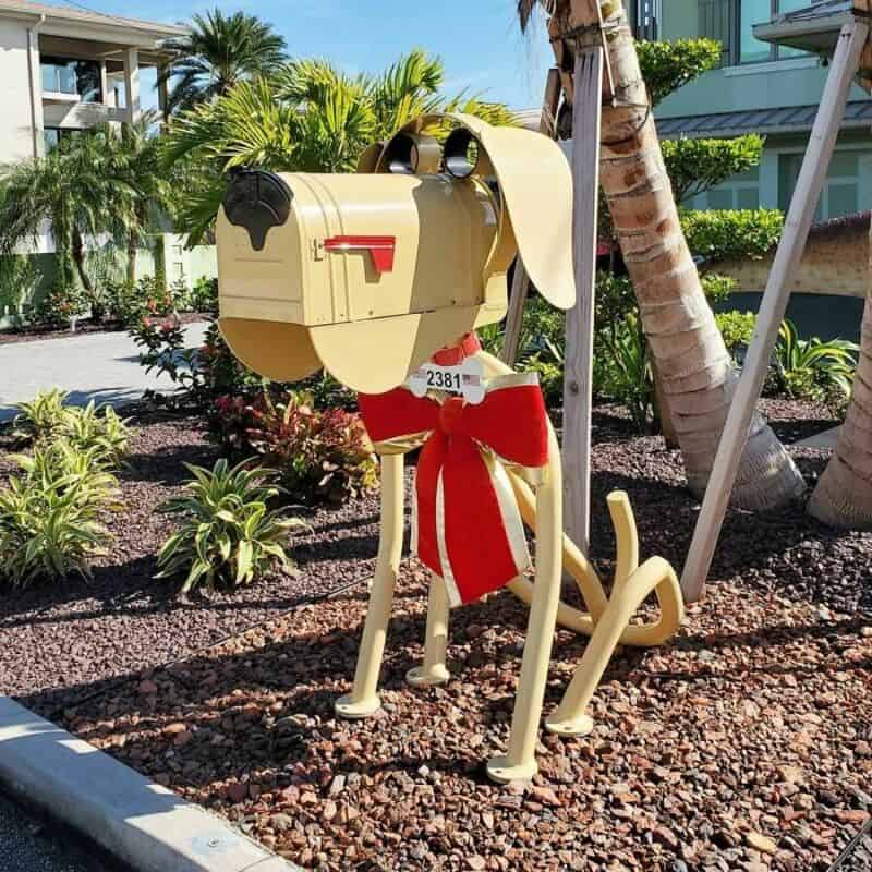 Dog-shaped mailbox with a red scarf, set among plants and trees.