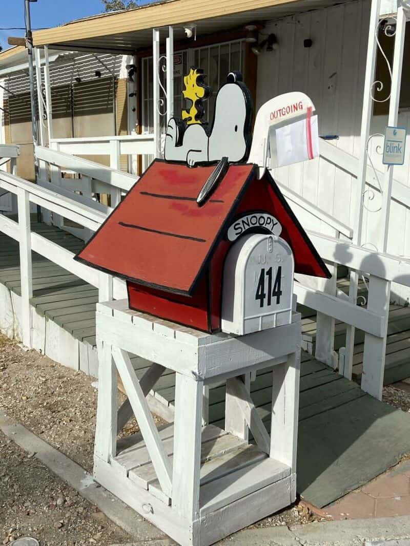 Snoopy mailbox with red roof, white body, and outgoing slot, on a white stand outside.
