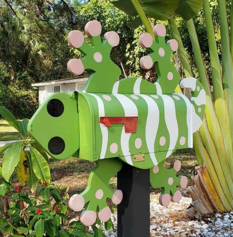 Colorful mailbox shaped like a chameleon with pink and green details, surrounded by tropical plants.