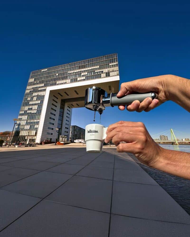 A hand holds an espresso machine pouring coffee into a cup in front of a modern building.