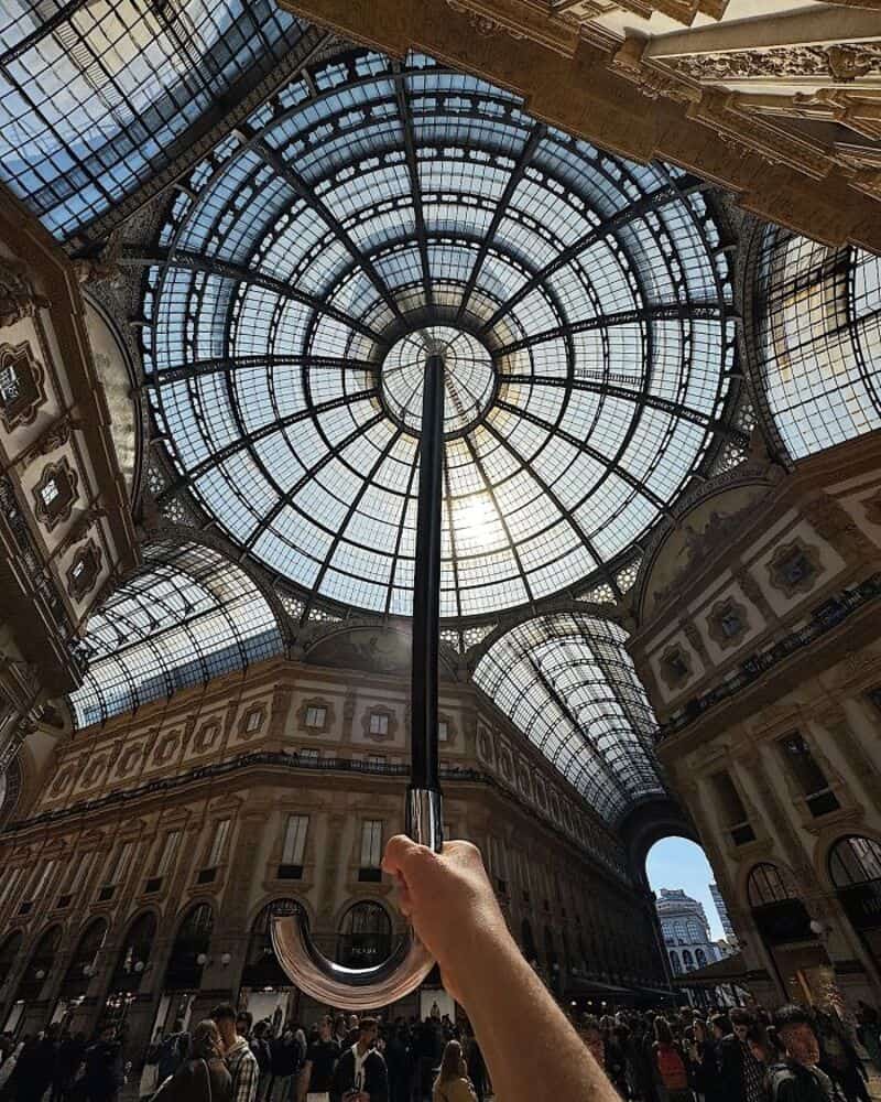 Hand holding a black umbrella under a glass-domed ceiling in a shopping gallery.