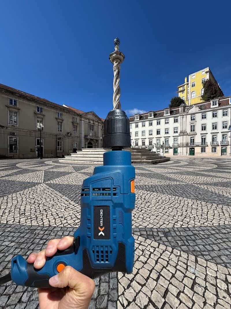 Power drill held in front of a monument in a city square under a clear blue sky.